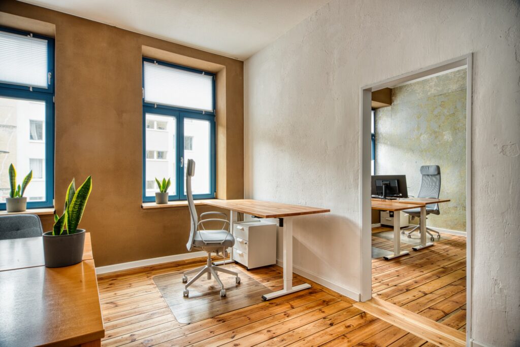 In einem Raum mit warmem Holzfußboden und reichlich Tageslicht stehen moderne, höhenverstellbare Büroschreibtische. Die natürliche Umgebung schafft eine inspirierende Arbeitsatmosphäre für maximale Produktivität.
